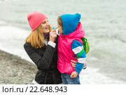 Мама обнимает маленькую дочку и весело трогает пальцем ее нос на берегу моря в холодную погоду. Стоковое фото, фотограф Иванов Алексей / Фотобанк Лори