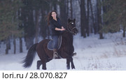 Beautiful longhaired woman riding a black horse through the snow in the forest, stallion prancing. Стоковое фото, фотограф Константин Шишкин / Фотобанк Лори