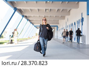 Portrait of young cheerful female traveler wearing casual clothes carrying heavy backpack and luggage at airport. Стоковое фото, фотограф Matej Kastelic / Фотобанк Лори