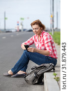 Mature Caucasian woman using smartphone for call for a taxi, sitting on roadside with backpack. Стоковое фото, фотограф Кекяляйнен Андрей / Фотобанк Лори