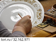 Мастер по росписи фарфора расписывает большую декоративную тарелку. Фарфоровая фабрика в Вербилках (2019 год). Редакционное фото, фотограф Наталья Николаева / Фотобанк Лори