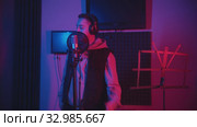 A man in headphones wearing a hoodie rapping through the pop-filter in the microphone - recording his track in the studio - music stand on the foreground. Стоковое видео, видеограф Константин Шишкин / Фотобанк Лори