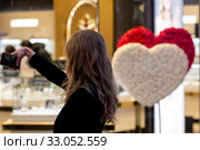 Красивая девушка делает селфи в торговом центре Метрополис на фоне витрины ювелирного магазина с оформлением приуроченным к Дню Святого Валентина в городе Москве, Россия (2020 год). Редакционное фото, фотограф Николай Винокуров / Фотобанк Лори
