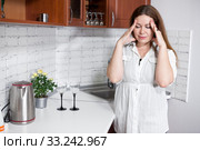 Stressed woman touching her head, standing in domestic kitchen with headache. Everyday routine duties of housewife. Стоковое фото, фотограф Кекяляйнен Андрей / Фотобанк Лори
