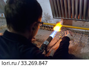 Молодой мужчина-стеклодув обрабатывает стеклянную заготовку при помощи газовой горелки в мастерской на фабрике ёлочных игрушек в поселке Крестцы Новгородской области (2019 год). Редакционное фото, фотограф Румянцева Наталия / Фотобанк Лори