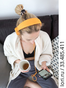 Stay at home. Social distancing. Woman at home relaxing on sofa couch drinking tea from white cup, listening to relaxing music, stay connected to friens and family via social networks on mobile. Стоковое фото, фотограф Matej Kastelic / Фотобанк Лори