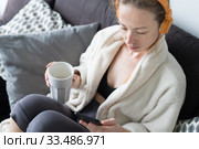 Stay at home. Social distancing. Woman at home relaxing on sofa couch drinking tea from white cup, listening to relaxing music, stay connected to friens and family via social networks on mobile. Стоковое фото, фотограф Matej Kastelic / Фотобанк Лори