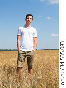 Full length portrait of teenager boy standing on agricultural field with ripe wheat, yellow meadow and blue sky, hands are in pockets of shorts, looking at camera. Стоковое фото, фотограф Кекяляйнен Андрей / Фотобанк Лори