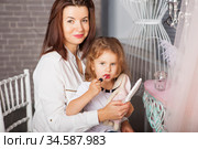 Lovely mother with daughter. Beautiful little lady looking at mirror and paints lips. fashion baby girl is doing makeup sitting on her mother's lap. Children Imitating Adults. Стоковое фото, фотограф Nataliia Zhekova / Фотобанк Лори