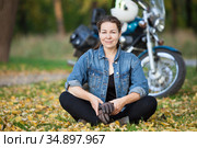 Attractive biker girl sits in the lotus position in front of stylish motorcycle at autumn park, smiling happy woman. Стоковое фото, фотограф Кекяляйнен Андрей / Фотобанк Лори
