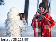 Caucasian woman taking picture with smartphone of her girlfriend for social network, girl wrapping red scarf and holding coffee mug at winter season. Стоковое фото, фотограф Кекяляйнен Андрей / Фотобанк Лори