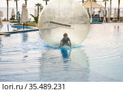 Little boy have fun inside big plastic balloon on the water of swimming pool on the summer resort. Kid inside big inflatable transparent ball running and having fun. Стоковое фото, фотограф Nataliia Zhekova / Фотобанк Лори