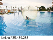 Little boy have fun inside big plastic balloon on the water of swimming pool on the summer resort. Kid inside big inflatable transparent ball running and having fun. Стоковое фото, фотограф Nataliia Zhekova / Фотобанк Лори