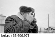 Портрет мужчина с фотоаппаратом, человек с камерой занимается фотографированием птицефермы, деревянные постройки и опора ЛЭП на заднем плане, Лорино, Чукотка, 1970. Редакционное фото, фотограф Варфоломей Тетерин / Фотобанк Лори