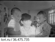 Взрослая женщина держит двух малышей на руках, она улыбается и смотрит на смеющихся детей, помещение с окнами на заднем плане, село Уэлен, Чукотка, 1970 год, полярная научная станция. Редакционное фото, фотограф Варфоломей Тетерин / Фотобанк Лори