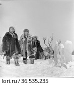 Оленевод Василий Кривошапкин и бригадир Н. Н. Текеянов позируют в традиционой Чукотской одежде, упряжки с северными оленями на заднем плане, Орджоникидзевский район, село Макарасы, март 1963. Редакционное фото, фотограф Варфоломей Тетерин / Фотобанк Лори