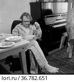 Человек курит трубку за обеденным столом, он что-то читает сидя в комнате рядом с роялем, взрослый мужчина в пижаме курит табак , Токарев и Морозов в гостях у Тетериных. Редакционное фото, фотограф Варфоломей Тетерин / Фотобанк Лори