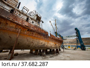 Mangystau, Kazakhstan - May 19, 2012: Old rusted ship on Caspian Sea, Bautino bay, ship-repairing yard. Marine Travelift Mobile Boat Hoist with belts on background. Grey stormy cloudy sky. Стоковое фото, фотограф Alexey Rezvykh / Фотобанк Лори