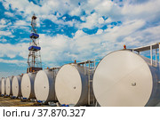 In the foreground are gray cylindrical tanks for storing oil and fuel reserves. Located horizontally in a row. In the background is a rig for drilling oil wells. Blue cloudy sky. Стоковое фото, фотограф Владимир Ендовицкий / Фотобанк Лори