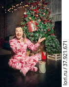 Cheerful funny man in pink sleepwear near decorated fir tree and throws up Christmas present. Стоковое фото, фотограф Darkbird77 / Фотобанк Лори