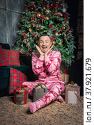 Cheerful surprised funny man in pink sleepwear sitting near decorated fir tree and Christmas present. Стоковое фото, фотограф Darkbird77 / Фотобанк Лори