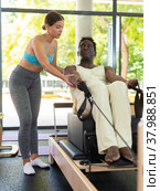 Positive activ female instructor controlling African American man during pulling stretch band sitting. Стоковое фото, фотограф Яков Филимонов / Фотобанк Лори