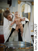 Infant baptism in Orthodox Church. Priest holds newborn child in his arms at baptismal font with Holy water. Baby is bathed in water. Sacrament rite of baptism. Редакционное фото, фотограф Aleksandr Golubev / Фотобанк Лори
