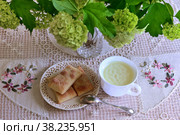 Домашняя кулинария. Сладкое печенье из слоеного теста на красивом блюдце и белая чашка с молочным киселем на фоне вязаной скатерти и вазы с цветами. Sweet homemade puff pastry cookies on a beautiful saucer and a white cup with milk jelly on the background of a knitted tablecloth and a vase with flowers. Стоковое фото, фотограф Владимир Литвинов / Фотобанк Лори