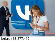 Россия. Санкт-Петербург. Молодые люди со смартфоном на фоне логотипа социальной сети ВКонтакте. Russia. Saint-Petersburg. Young people with a smartphone on the background of the VKontakte social network logo. (2022 год). Редакционное фото, фотограф Алексей Смышляев / Фотобанк Лори
