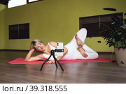 Attractive blonde woman, yoga teacher, streaming with a smartphone to her audience, lies on a mat and performs a lotus position, has an online workout in a studio, communicates with clients. Стоковое фото, фотограф Сергей Чайко / Фотобанк Лори