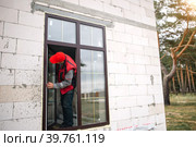 The window installer adjusts the hinges and checks new windows in the cottage under construction. Ordering windows with golden layout and brown lamination for a private house. A worker in uniform. Стоковое фото, фотограф Ольга Симонова / Фотобанк Лори