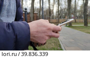 Молодой человек сидит на скамейке в городском парке, набирает текстовое сообщение или  играет в игру в мобильном телефоне. Смартфон в мужских руках. Мужчина делает онлайн покупки на сайте маркетплейса. A young man is sitting on a bench in a city park, typing a text message or playing a game on a mobile phone. The smartphone is in men's hands. A man makes online purchases on the marketplace website. Стоковое видео, видеограф Ирина / Фотобанк Лори