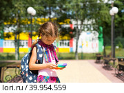Little girl with a backpack and in a school uniform in the school yard plays pop it toy. Back to school, September 1. The pupil relaxes after lessons. Primary education, elementary class for student. Стоковое фото, фотограф Ольга Симонова / Фотобанк Лори