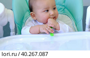 A hungry baby is gnawing on a plastic spoon at the table on a high chair. Teething, whims, itchy gums, introduction of complementary foods. Стоковое видео, видеограф Ольга Симонова / Фотобанк Лори