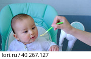 Mom feeds the baby with a spoon of vegetable puree at the children's feeding table. Baby's appetite, healthy nutrition, introduction of complementary foods. Copyspace, mock up. Стоковое видео, видеограф Ольга Симонова / Фотобанк Лори