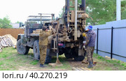 Team of workers with drilling rig on car are drilling artesian well for water in ground. Insertion of metal casing pipe into ground, installation of individual drinking supply, June 28, 2022, Russia, Palaces. Редакционное видео, видеограф Ольга Симонова / Фотобанк Лори