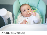 A hungry baby is gnawing on a plastic spoon at the table on a high chair. Teething, whims, itchy gums, introduction of complementary foods. Стоковое фото, фотограф Ольга Симонова / Фотобанк Лори