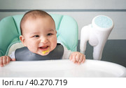 A dirty, joyful baby at the table on a feeding chair got his mouth dirty in vegetable puree. Introduction of complementary foods, the child likes to eat. Portrait close-up. Стоковое фото, фотограф Ольга Симонова / Фотобанк Лори