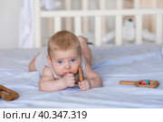 A charming boy in a bright bedroom. A newborn baby is lying on the bed and gnawing on toys. Teeth are teething. The baby's gums are itching. A child of 4 months looks into the camera. Family, pregnancy and childbirth. Стоковое фото, фотограф Photojuli / Фотобанк Лори