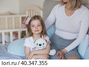 A happy young mother combs her adorable little daughter's hair while sitting in bed in the morning. The girl smiles. Family and care. Стоковое фото, фотограф Photojuli / Фотобанк Лори