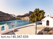 Greece. Rhodes. View of traditional Greek Christian white Church of Saint Paul on hill above St. Paul's bay with blue water against background of columns of ancient Acropolis of Lindos on mountain (2016 год). Стоковое фото, фотограф Виктория Катьянова / Фотобанк Лори