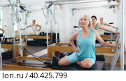 Svelte elderly woman performing set of pilates exercises on reformer during group workout. Active lifestyle and wellness concept of seniors. Стоковое видео, видеограф Яков Филимонов / Фотобанк Лори
