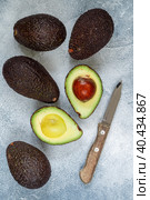 Whole and cut in half ripe organic avocados and a knife on a grey background. The Haas Avocado. Source of vitamins , trace elements , beta-carotene and omega-3 fatty acids . Healthy food, diet. Стоковое фото, фотограф Виктория Панченко / Фотобанк Лори