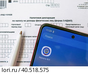 Телефон с электронным приложением налоговой службы "Налоги ФЛ" лежит на бланке налоговой декларации 3-НДФЛ. Оформление социального и имущественного вычета через интернет (2023 год). Редакционное фото, фотограф Татьяна Т / Фотобанк Лори