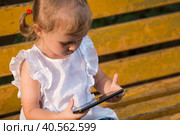 Cute little girl of 3 years old with a smartphone in her hands on a bench. A small child is playing on the phone. The harm of gadgets for children. Protect the child from the smartphone. Kids and gadgets. Стоковое фото, фотограф Photojuli / Фотобанк Лори