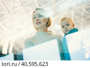 Thoughtful young mother looking trough window holding his infant baby boy child while waiting to board an airplane at airport terminal departure gates. Travel with baby concept. Стоковое фото, фотограф Matej Kastelic / Фотобанк Лори