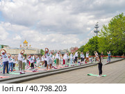 Пенсионеры, участники спортивного праздника программы "Московское долголетие" в фирменных московских футболках и шапочках выполняют гимнастику под руководством опытного тренера в парке Музеон на фоне храма Христа Спасителя, голубого неба и кучевых облаков (2023 год). Редакционное фото, фотограф Сергей Никифоров-Гордеев / Фотобанк Лори