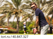 Young man golfer hitting golf ball on course in sportswear and sunglasses, teeing golf in club training at palm trees background. Healthy sports lifestyle and leisure activity concept. Copy text space. Стоковое фото, фотограф Aleksandr Golubev / Фотобанк Лори