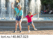 Two happy girls sisters in a bright summer t-shirt with colored African braids in the hair have fun by a city fountain. The children have a rest near a fountain during a heat wave. Стоковое фото, фотограф Ирина Михайличенко / Фотобанк Лори