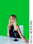 Cute young woman broadcaster sitting at desk with microphone at green backdrop, looking at camera. Portrait of female broadcaster posing in television studio. Tv production concept. Copy ad text space. Стоковое фото, фотограф Aleksandr Golubev / Фотобанк Лори